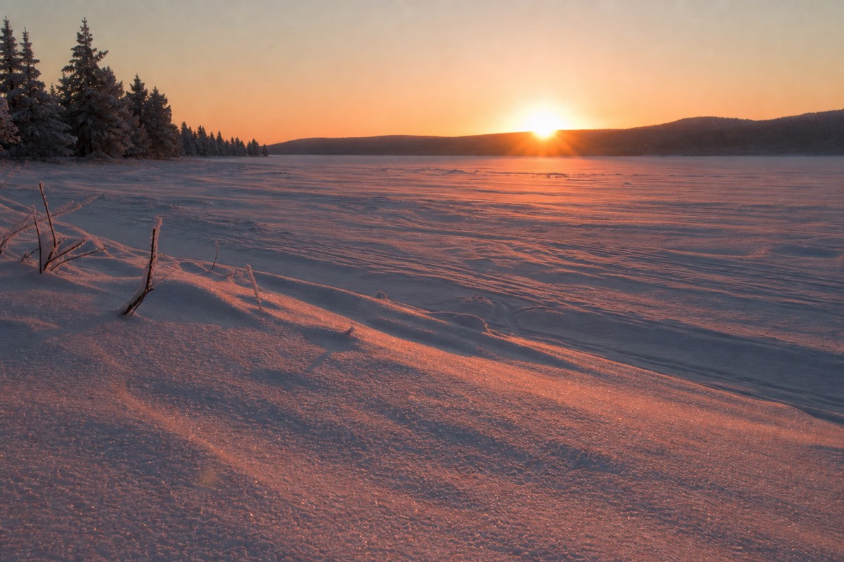 A low winter sun just above the horizon over a frozen Lapland lake, long pink light across snow