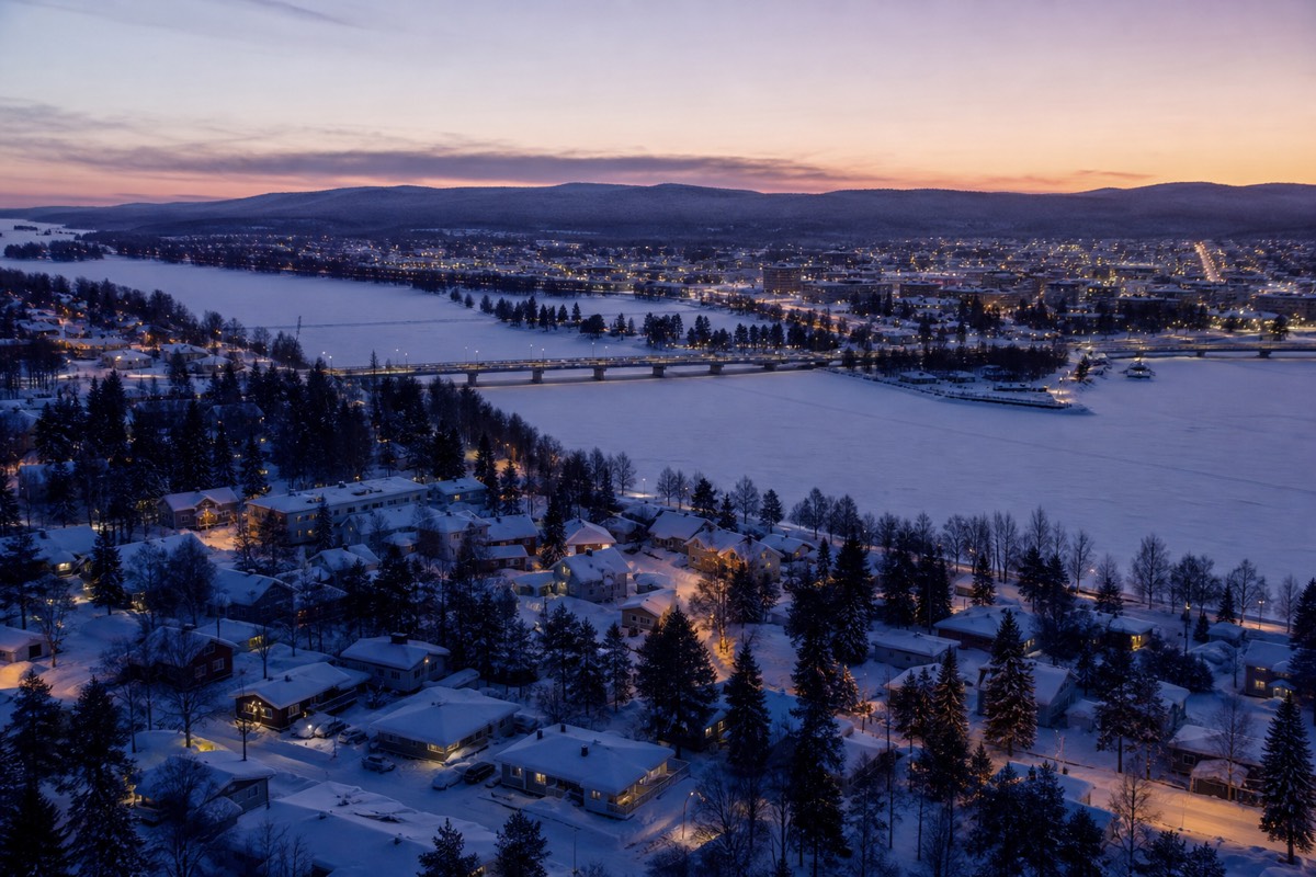 An aerial-style wide view of a snow-covered Lapland town beside a frozen river at dusk