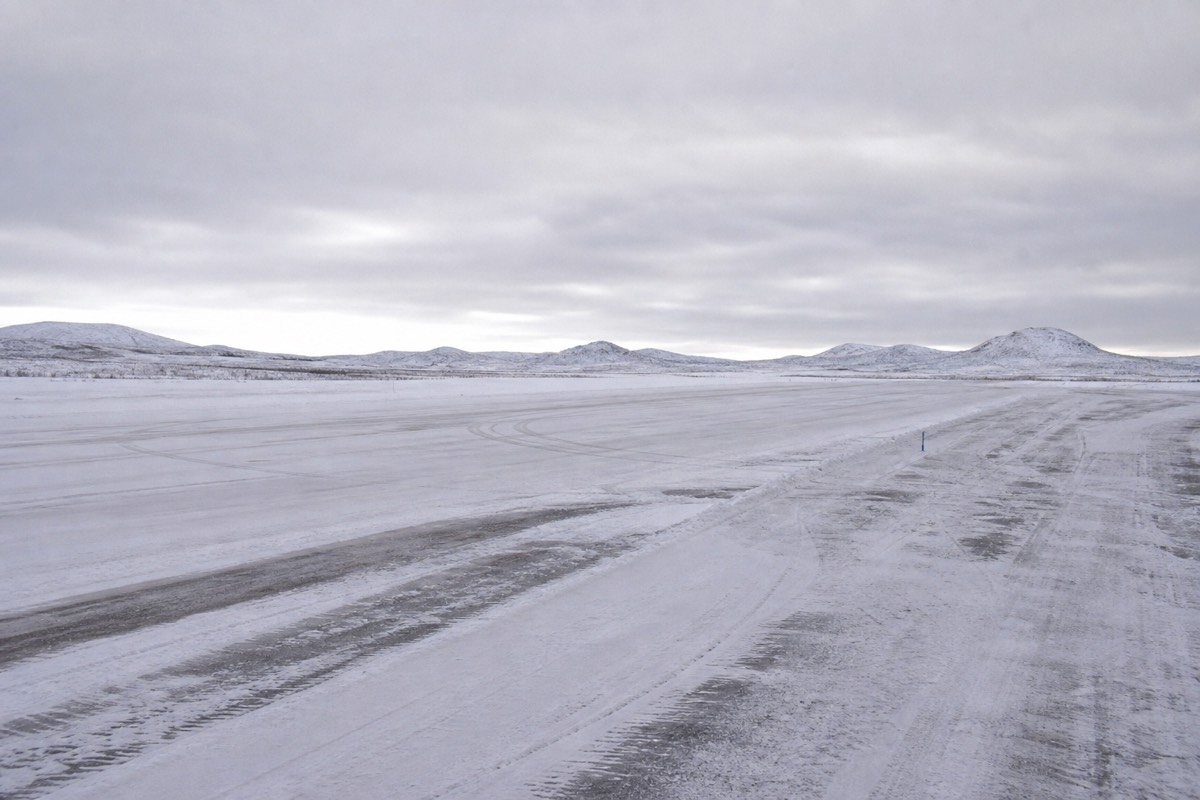 A snow-covered runway and taxiway at a small Arctic airport under heavy grey winter sky