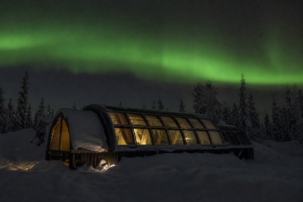 The curved glass roof of an igloo cabin seen from outside at night, faint green aurora above, snow around it