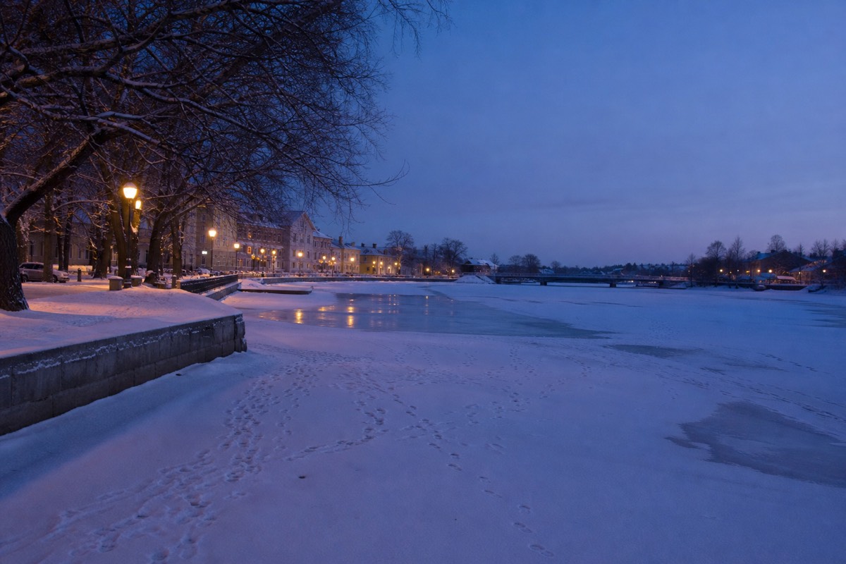 A frozen river running through a snowy Finnish town at blue hour, footprints in fresh snow