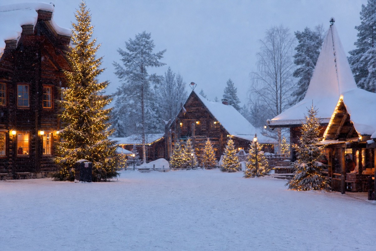 A snowy Nordic village square with wooden cabins and festive lights under falling snow