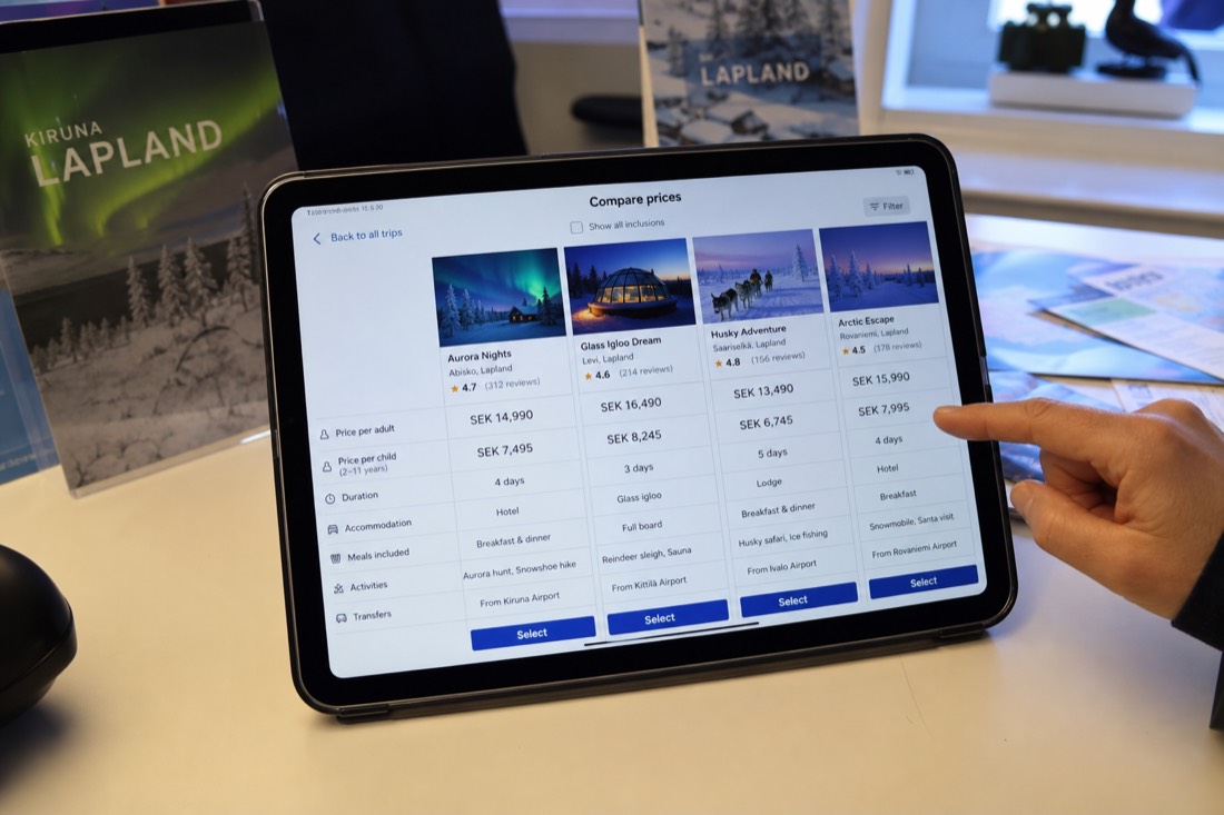 A digital price comparison chart displayed on a tablet in a travel planning office setting, winter Lapland photos visible