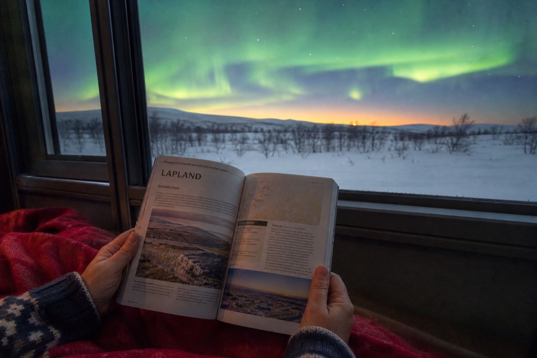A person holding open a Lapland travel guidebook, sitting by a window overlooking snowy landscape and distant aurora
