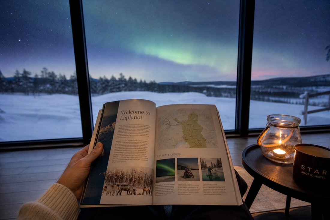 Una persona che sfoglia una guida turistica della Lapponia, seduta vicino a una finestra con vista su un paesaggio innevato e un’aurora boreale in lontananza