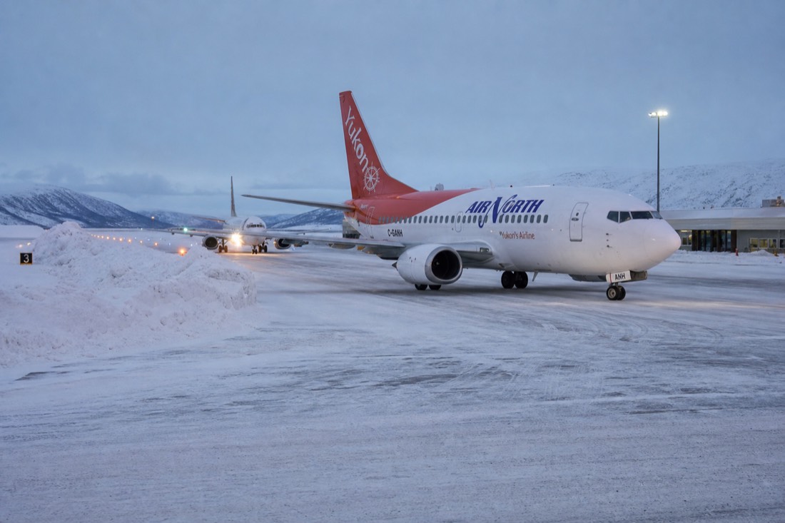 A small regional airport apron in deep Arctic snow, runway lights glowing, snowbanks, aircraft waiting