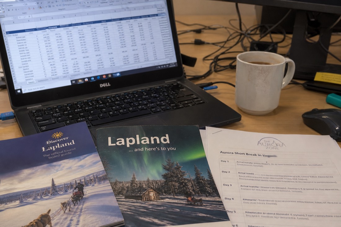 A spreadsheet with travel costs and pricing data on a desk with Lapland travel brochures and a coffee cup
