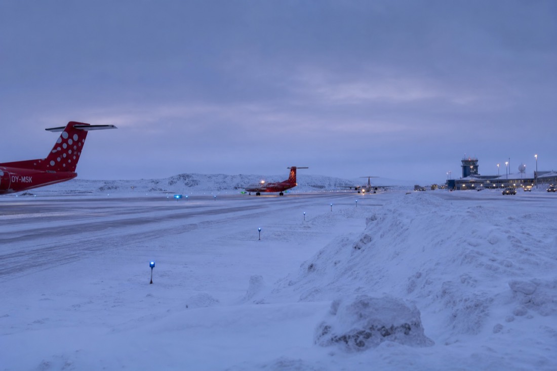A small regional airport apron in deep Arctic snow, runway lights glowing, snowbanks, aircraft waiting