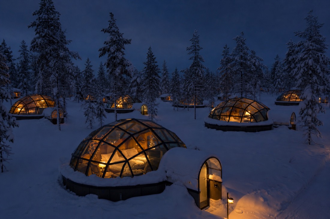 A cluster of glass-ceiling igloo cabins nestled in snow among trees, warm interior lighting, dark Lapland night sky above
