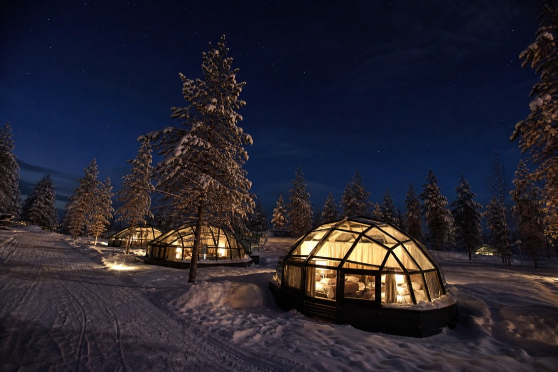 A cluster of glass-ceiling igloo cabins nestled in snow among trees, warm interior lighting, dark Lapland night sky above