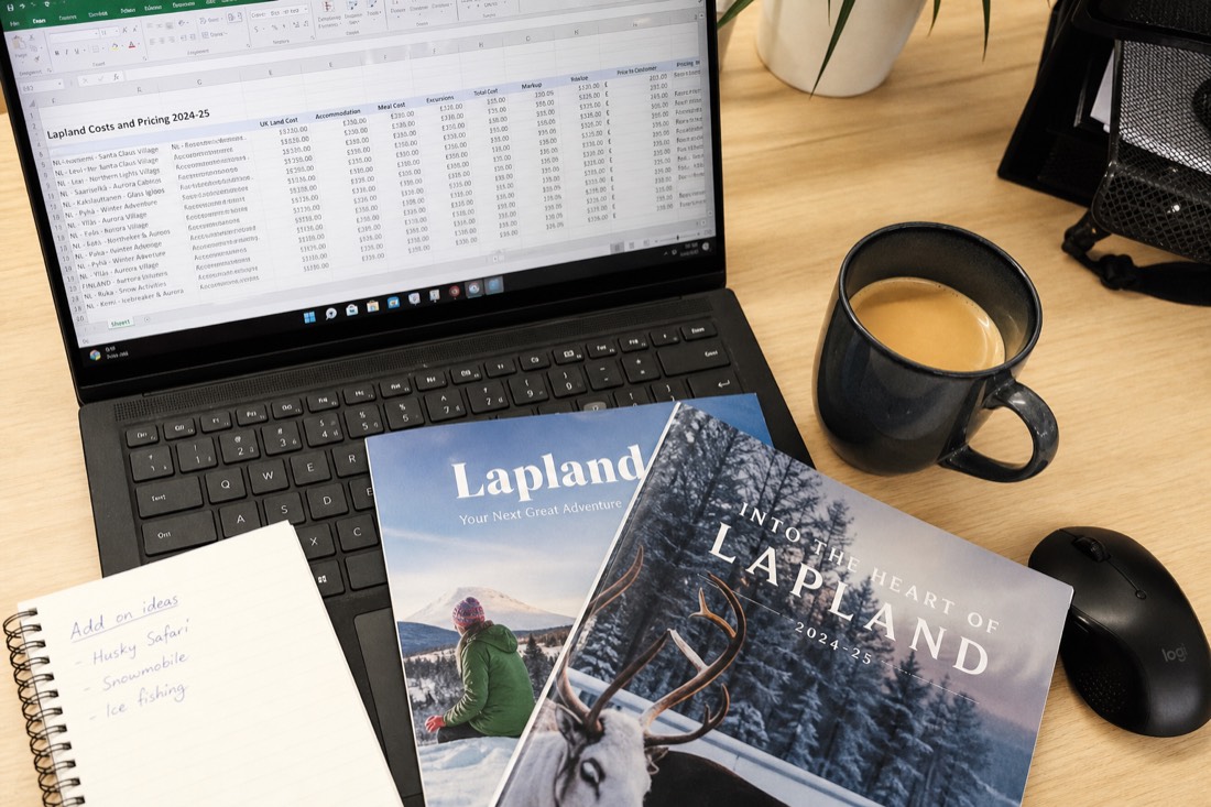 A spreadsheet with travel costs and pricing data on a desk with Lapland travel brochures and a coffee cup