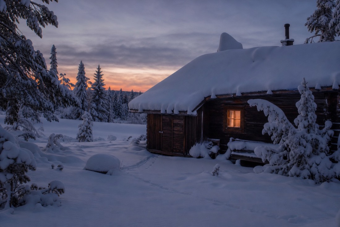 Ein hölzernes Lappland-Haus unter dickem Schnee in der Dämmerung, ein Fenster warm beleuchtet