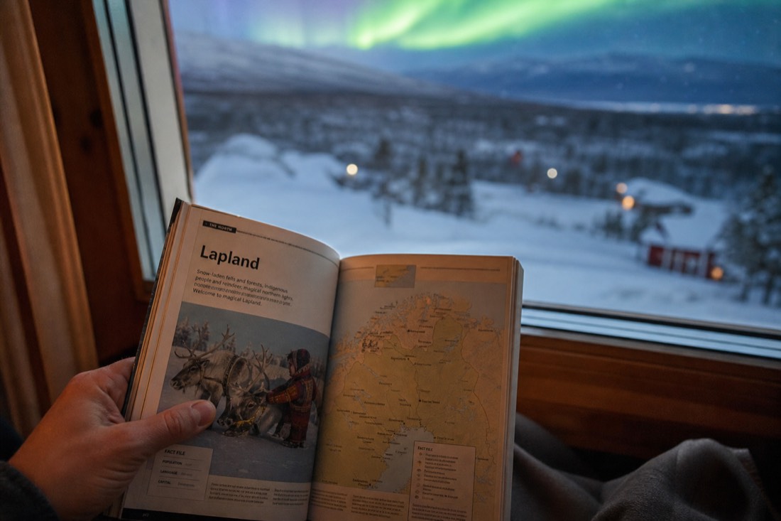 A person holding open a Lapland travel guidebook, sitting by a window overlooking snowy landscape and distant aurora