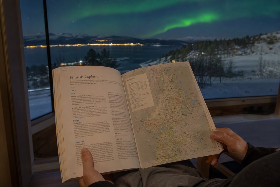 A person holding open a Lapland travel guidebook, sitting by a window overlooking snowy landscape and distant aurora