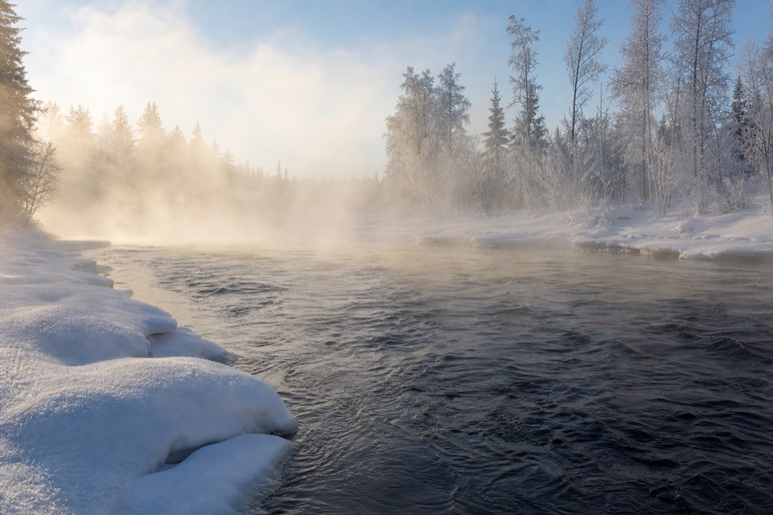 Steam rising off an open stretch of river in deep winter, snowbanks either side