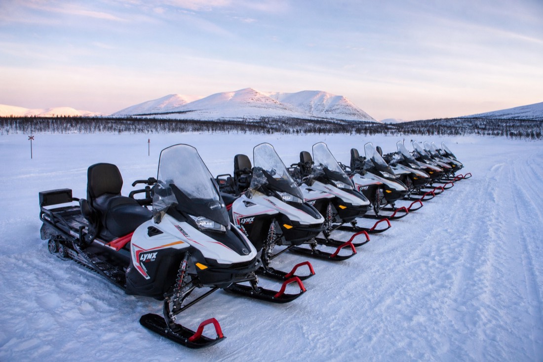 A group of modern snowmobiles parked in formation on deep snow, mountains visible in distance, Lapland winter landscape
