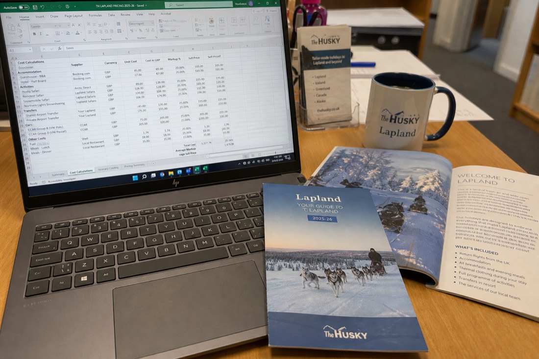 A spreadsheet with travel costs and pricing data on a desk with Lapland travel brochures and a coffee cup