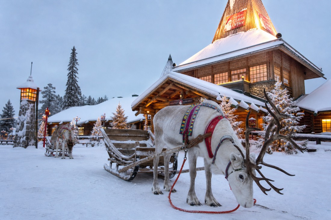 Santa Claus Village in Rovaniemi with reindeer outside wooden buildings in deep snow, warm lit windows, Arctic setting