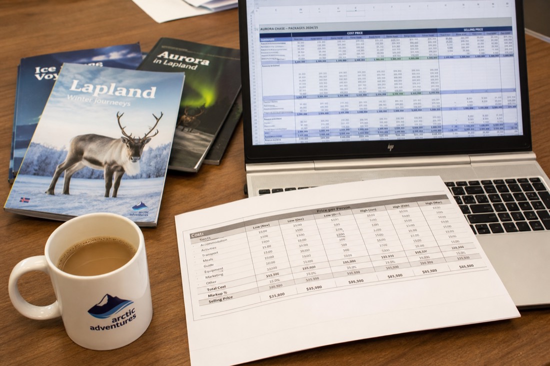 A spreadsheet with travel costs and pricing data on a desk with Lapland travel brochures and a coffee cup