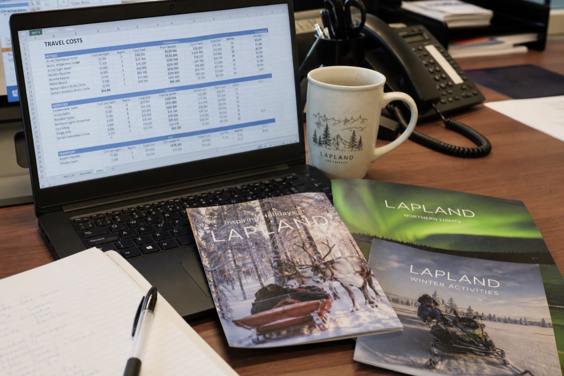 A spreadsheet with travel costs and pricing data on a desk with Lapland travel brochures and a coffee cup