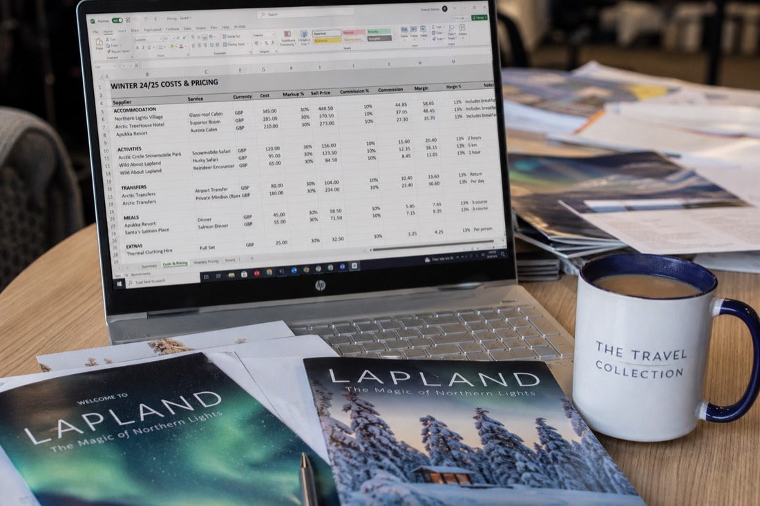 A spreadsheet with travel costs and pricing data on a desk with Lapland travel brochures and a coffee cup