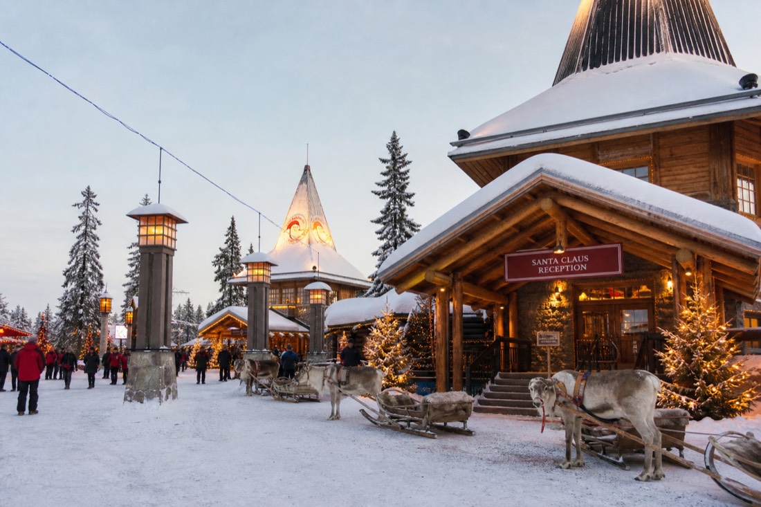 Santa Claus Village in Rovaniemi with reindeer outside wooden buildings in deep snow, warm lit windows, Arctic setting