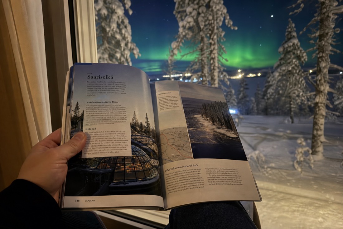 A person holding open a Lapland travel guidebook, sitting by a window overlooking snowy landscape and distant aurora
