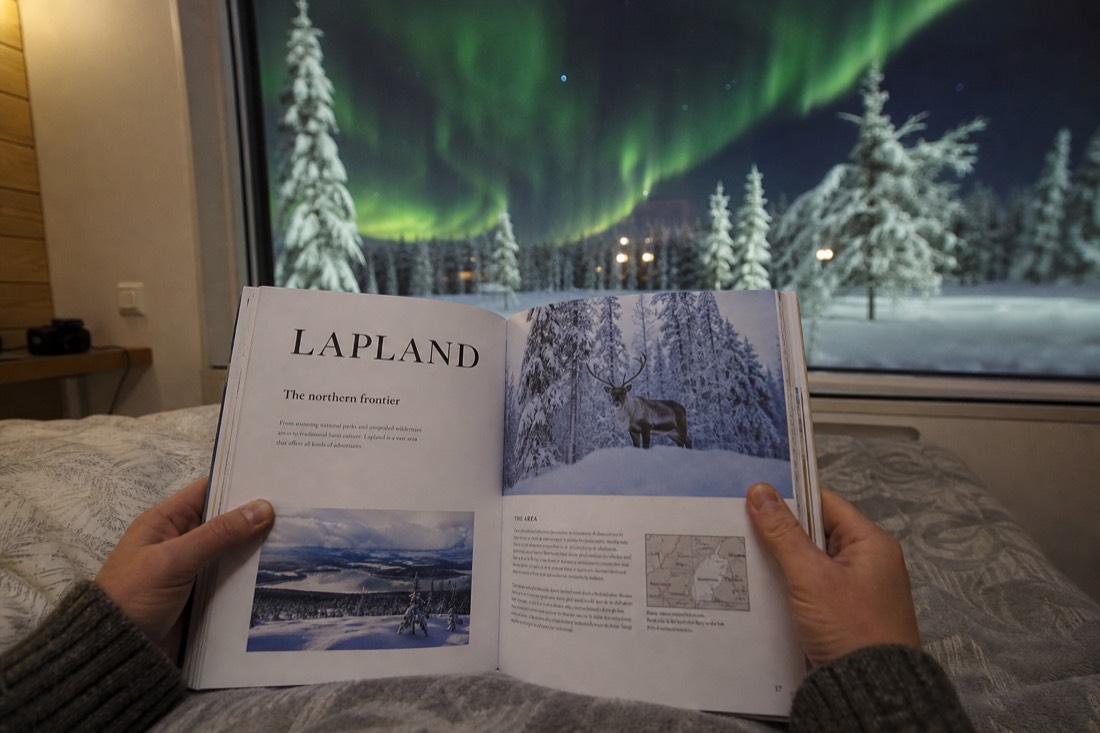 A person holding open a Lapland travel guidebook, sitting by a window overlooking snowy landscape and distant aurora