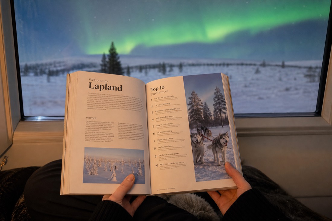 A person holding open a Lapland travel guidebook, sitting by a window overlooking snowy landscape and distant aurora