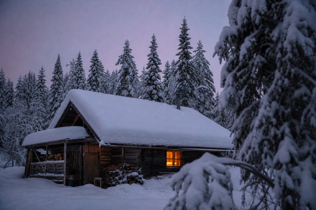 Ein hölzernes Lappland-Haus unter dickem Schnee in der Dämmerung, ein Fenster warm beleuchtet