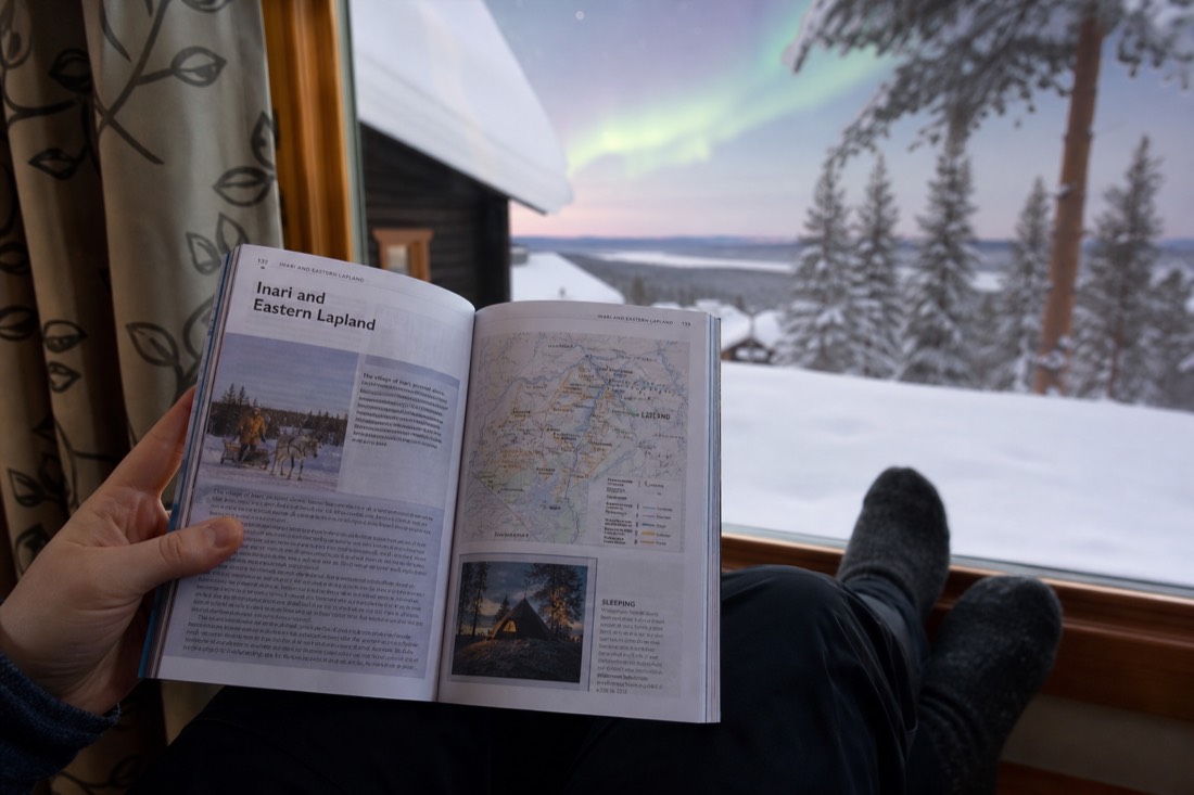 A person holding open a Lapland travel guidebook, sitting by a window overlooking snowy landscape and distant aurora