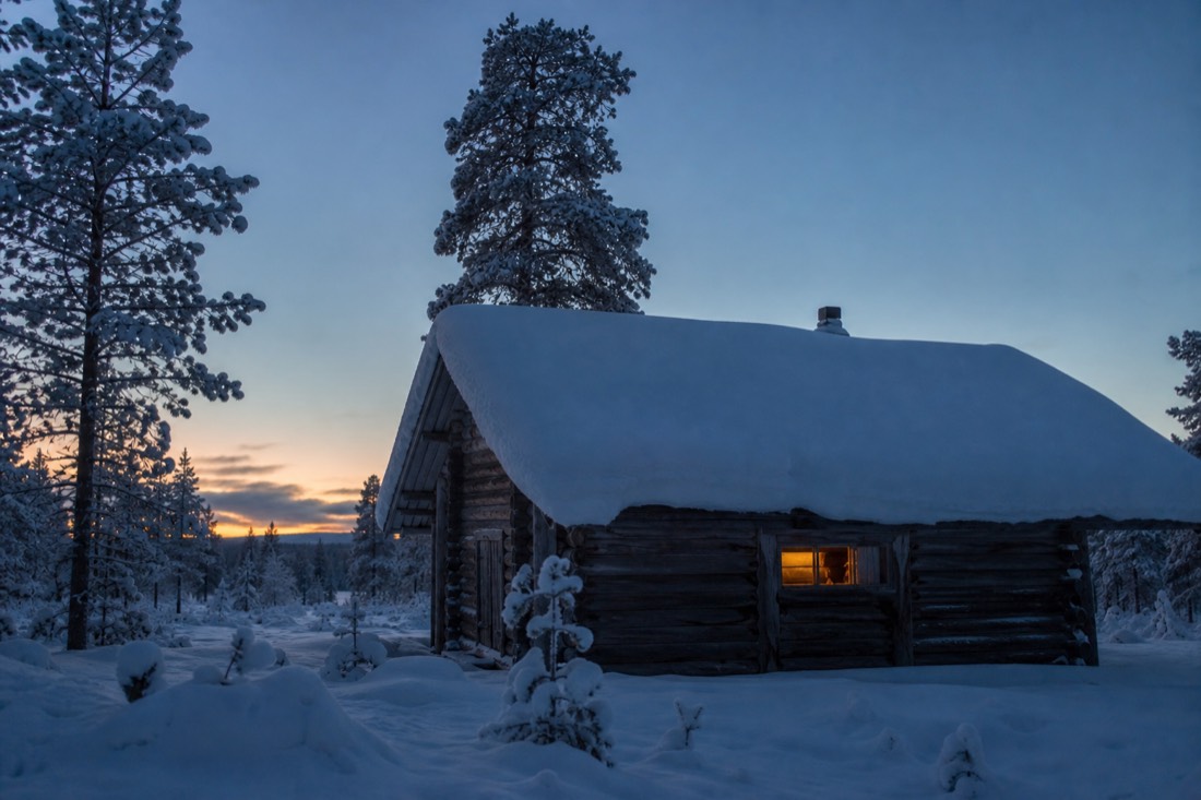 Una baita in legno della Lapponia coperta di neve al tramonto, con una finestra illuminata