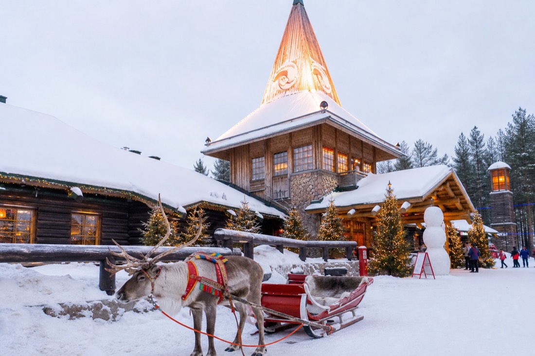 Santa Claus Village in Rovaniemi with reindeer outside wooden buildings in deep snow, warm lit windows, Arctic setting