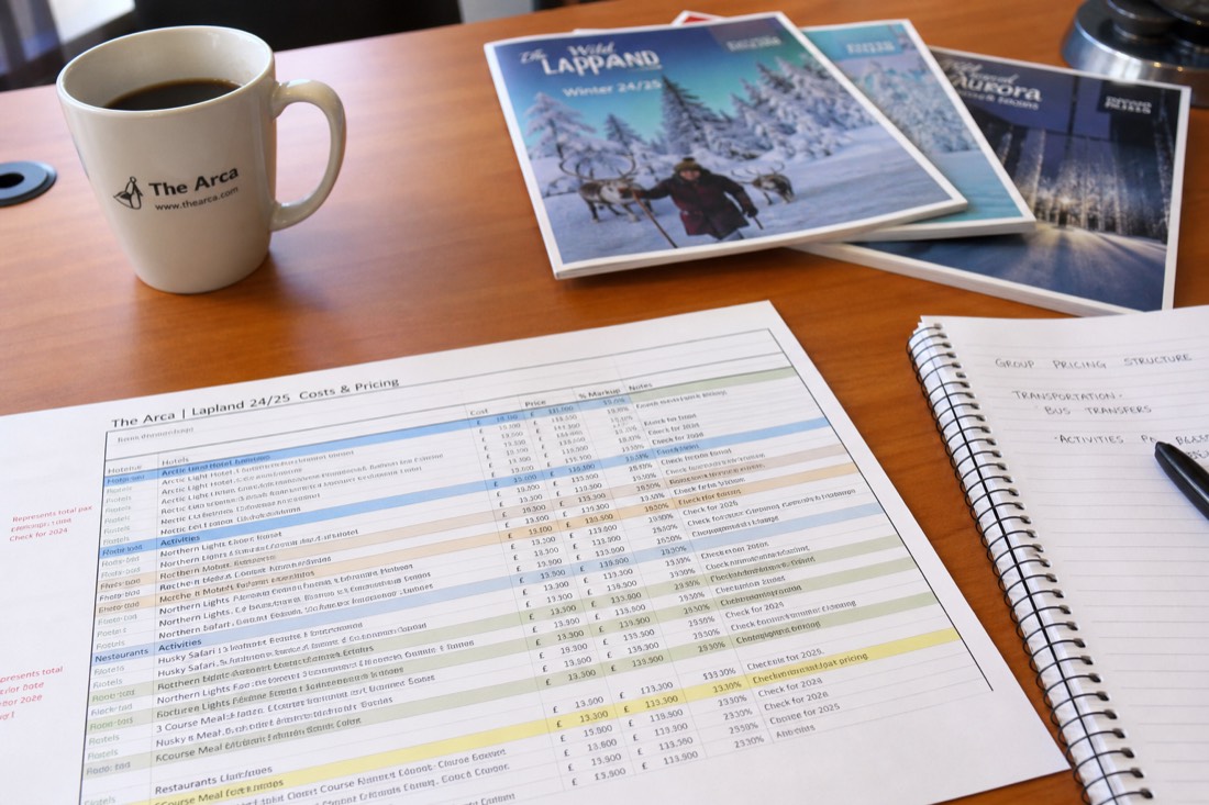 A spreadsheet with travel costs and pricing data on a desk with Lapland travel brochures and a coffee cup