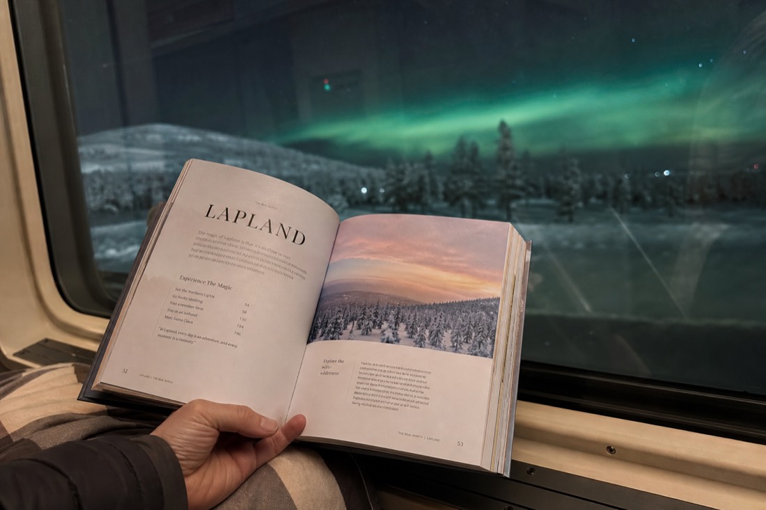 A person holding open a Lapland travel guidebook, sitting by a window overlooking snowy landscape and distant aurora