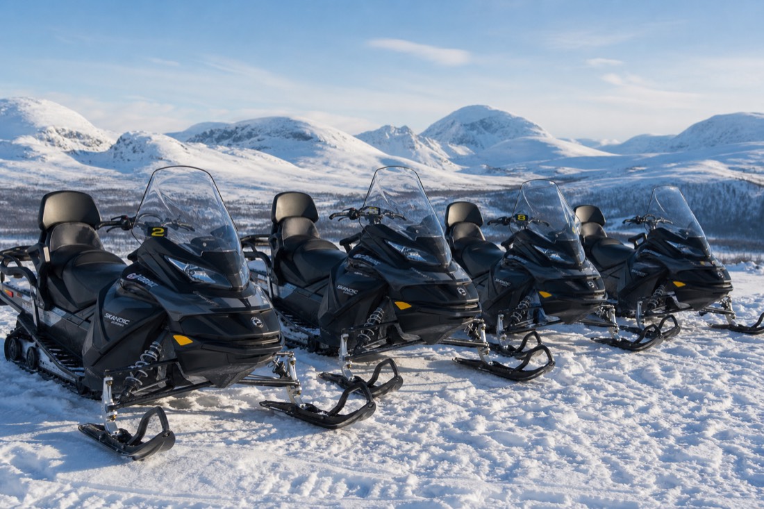 A group of modern snowmobiles parked in formation on deep snow, mountains visible in distance, Lapland winter landscape