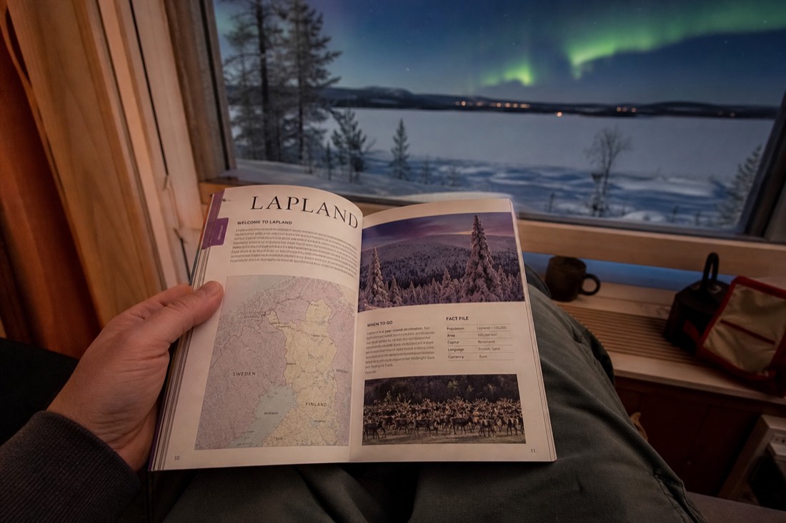 A person holding open a Lapland travel guidebook, sitting by a window overlooking snowy landscape and distant aurora