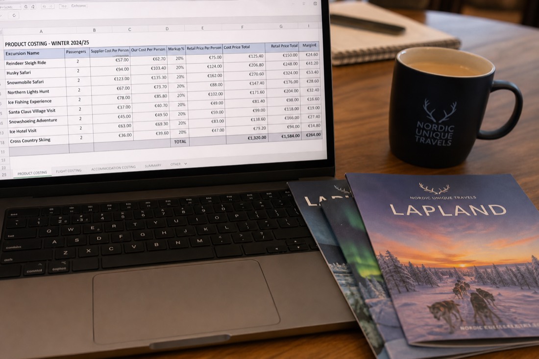 A spreadsheet with travel costs and pricing data on a desk with Lapland travel brochures and a coffee cup