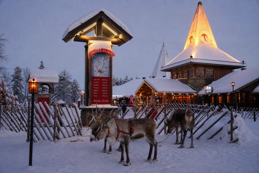 Santa Claus Village in Rovaniemi with reindeer outside wooden buildings in deep snow, warm lit windows, Arctic setting