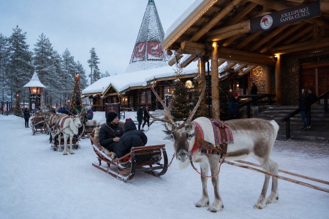 Santa Claus Village in Rovaniemi with reindeer outside wooden buildings in deep snow, warm lit windows, Arctic setting