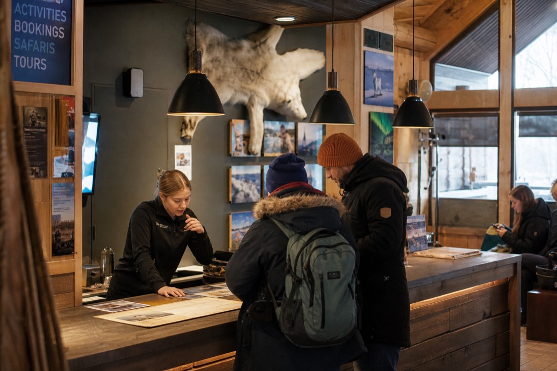 Customer service desk at a modern Lapland activity booking center, staff helping tourists, real wooden interior, warm lighting