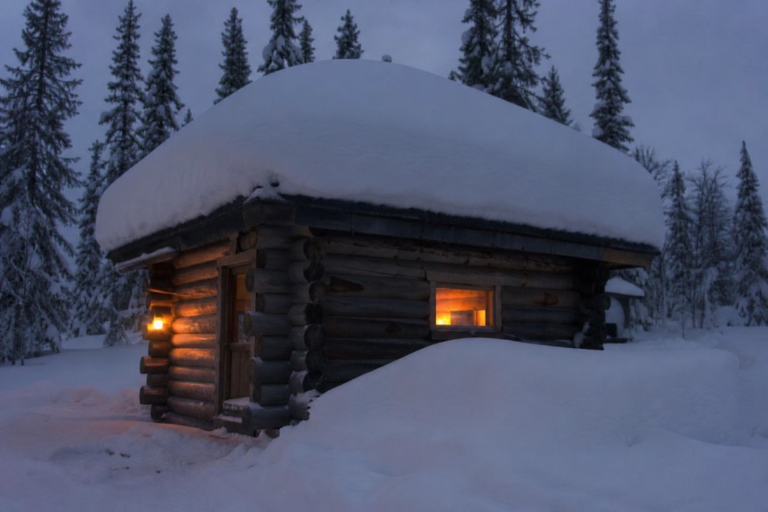 A wooden Lapland cabin under thick snow at dusk, one window lit warm