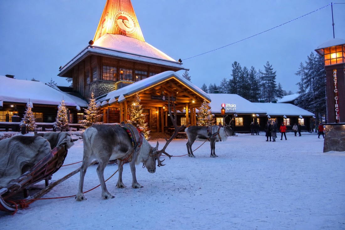 Santa Claus Village in Rovaniemi with reindeer outside wooden buildings in deep snow, warm lit windows, Arctic setting